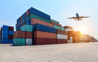 Airplane flying over stacked shipping containers at a port logistics yard during sunrise