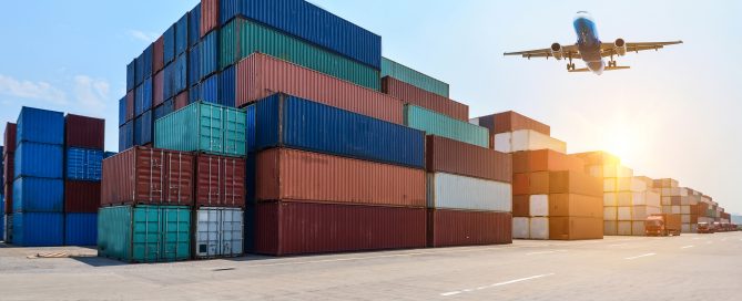 Airplane flying over stacked shipping containers at a port logistics yard during sunrise